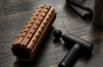 Wooden foam roller and black massage gun side by side on dark gym floor with towel and water bottle