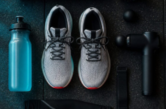 Running shoes with foam roller, massage gun, compression sleeve and electrolyte bottle in flat lay on dark gym floor