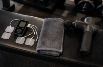TENS unit with electrode pads and massage gun side by side on gym bench