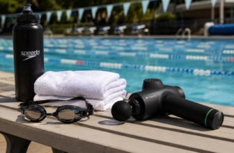 Massage gun, swim goggles, towel and water bottle on a pool deck bench