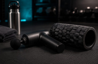 Black massage gun next to a dark foam roller on a gym floor mat